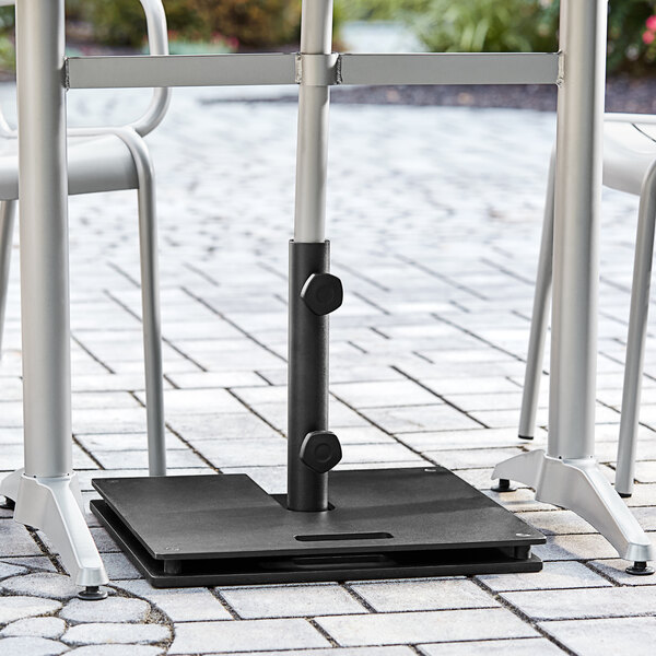 A Lancaster Table & Seating black steel umbrella base with wheels under a table with chairs on a brick surface.