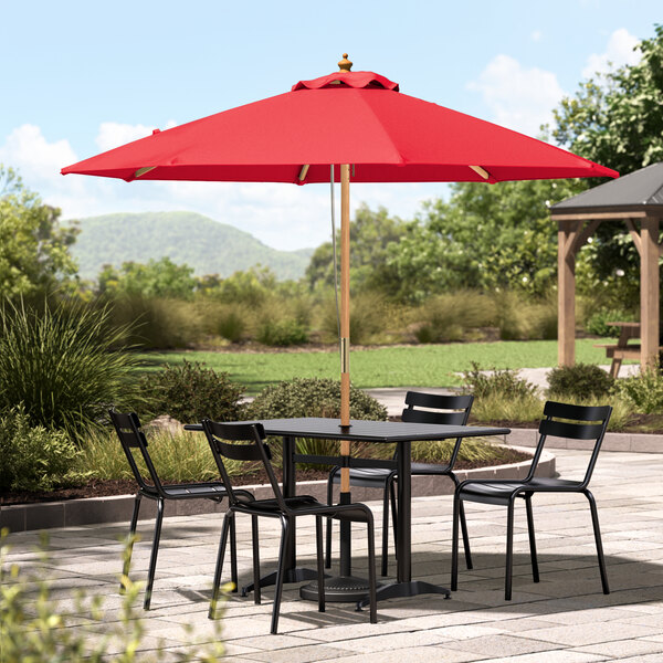 A Lancaster Table & Seating strawberry umbrella with a wooden pole over a table and chairs on a patio.