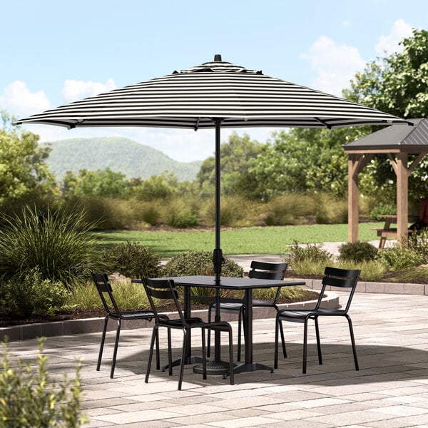 A table with black chairs under a black and white striped Lancaster Table and Seating umbrella on a patio.