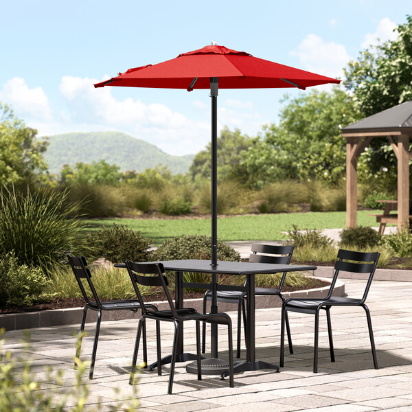 A black table and chairs under a Lancaster Table & Seating red umbrella on a stone patio.
