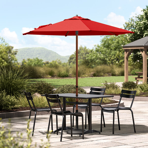 A wooden table and chairs under a red Lancaster Table & Seating umbrella on a patio.