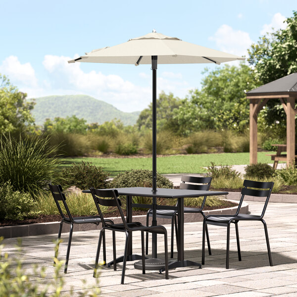 A round ivory table with black chairs and a Lancaster Table & Seating black umbrella on a stone patio.