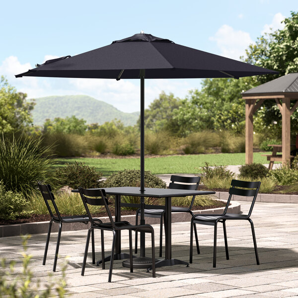 A black table with black metal chairs under a navy umbrella on a patio.