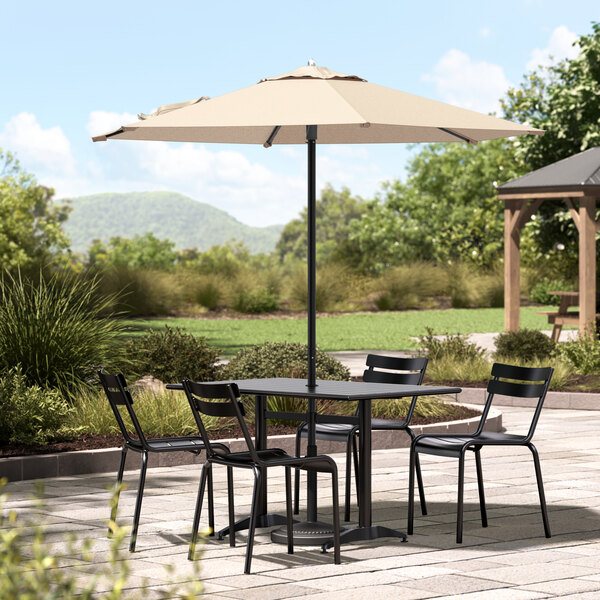 A Lancaster Table & Seating black aluminum umbrella over a table with chairs on a stone patio.