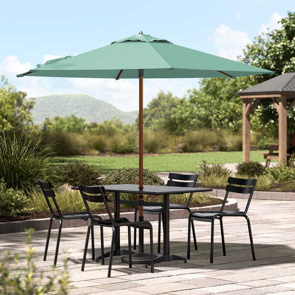 A wooden table and black chairs under a green Lancaster Table & Seating umbrella on a stone patio.