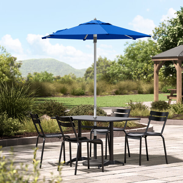 A table with black chairs and a Lancaster Table & Seating blue umbrella on a stone patio.
