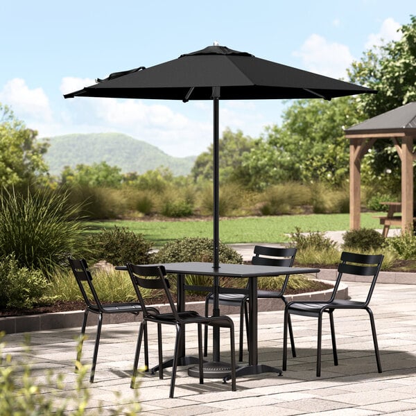 A black Lancaster Table & Seating umbrella over a table and chairs on a stone patio.