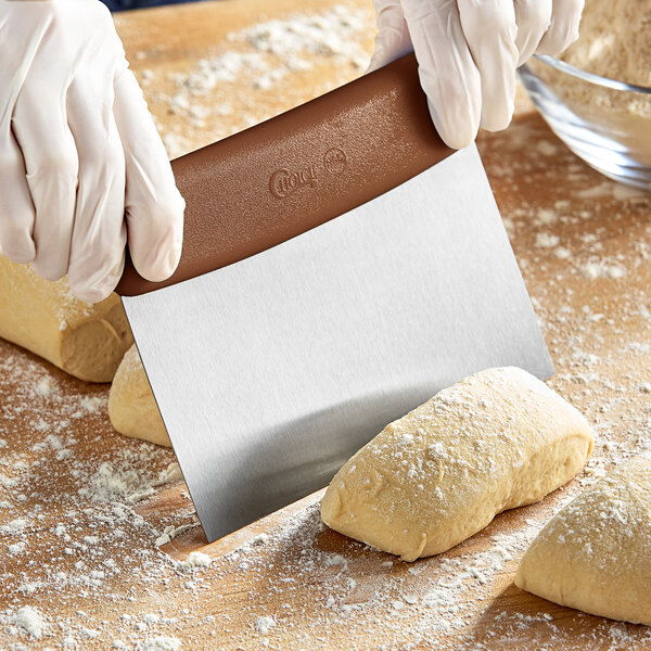 A person using a Choice stainless steel dough cutter with a brown handle to cut dough.