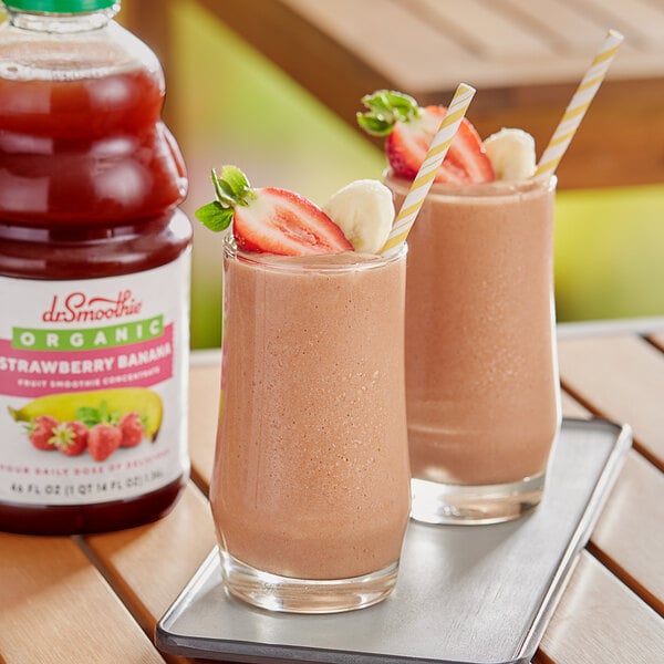 Two glasses of pink Dr. Smoothie Organic Strawberry Banana smoothies with straws and fruit on a tray.