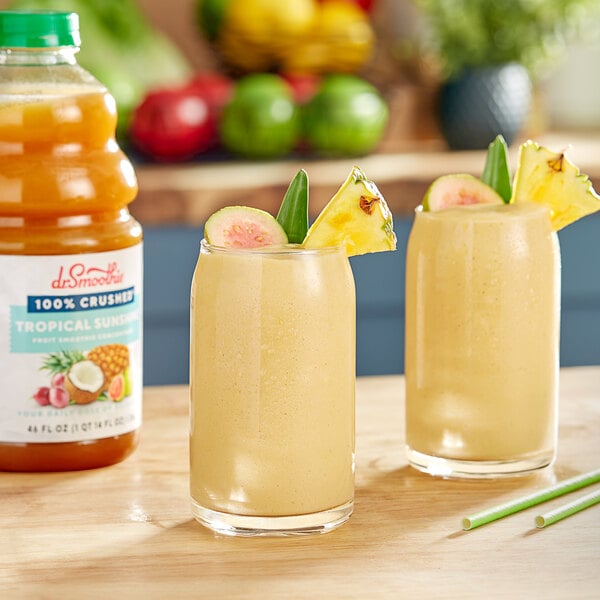 Two glasses of yellow Dr. Smoothie Tropical Sunshine smoothie with pineapple garnish on a table.