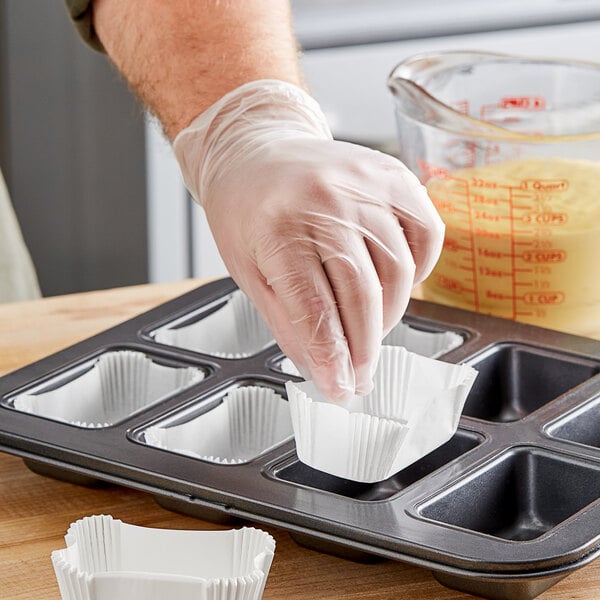 A person in gloves putting a white fluted oblong loaf liner into a black muffin pan.