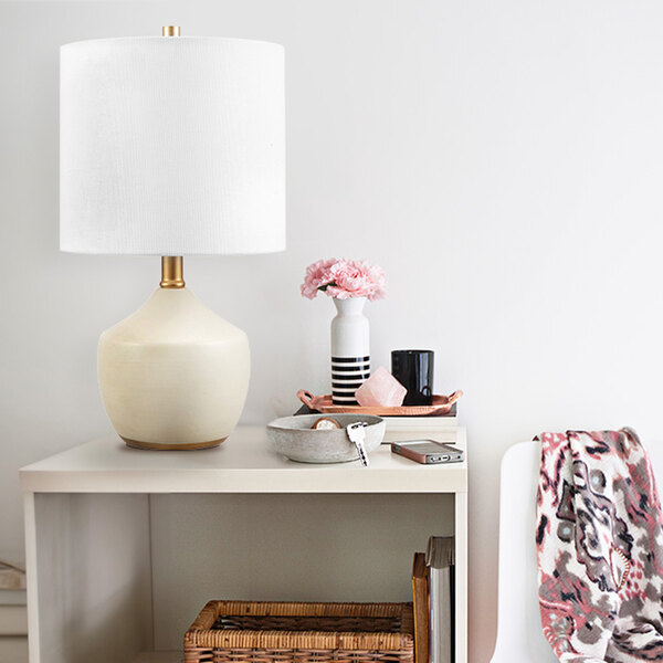 A white ceramic table lamp with brass accents on a white table with a vase of flowers.