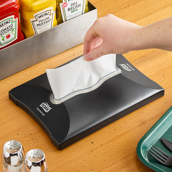 A hand putting a napkin in a black Tork interfold napkin dispenser on a counter.