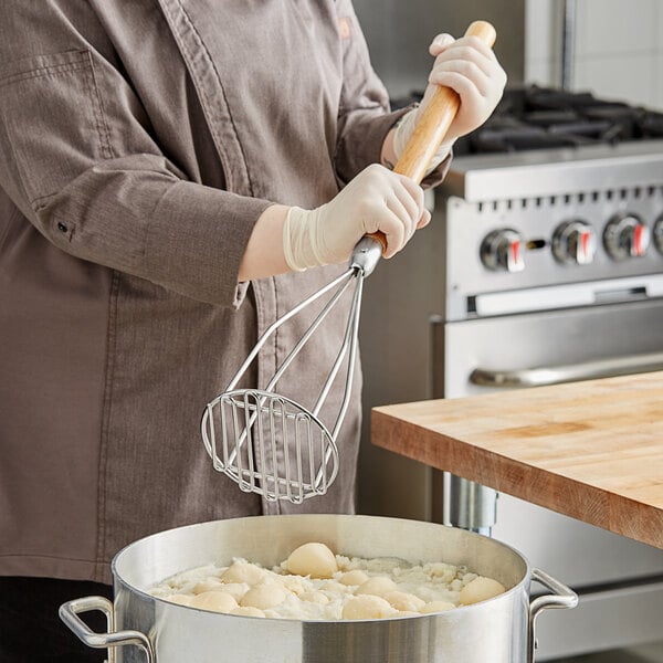 A woman in a chef's uniform using a wood-handled round potato masher to stir a pot of food.