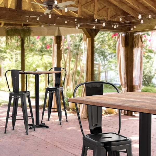 A Lancaster Table & Seating Excalibur bar height table with 2 black metal chairs with wooden seats on a covered patio.