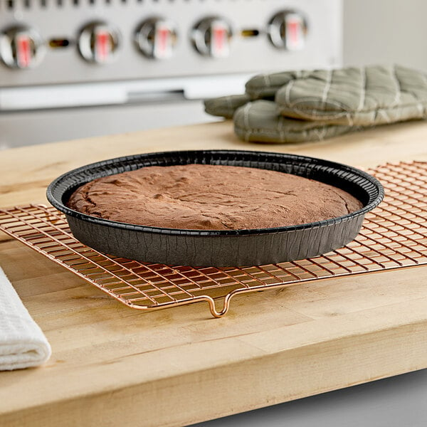 A brown cake in a Solut round black paperboard pan on a cooling rack.