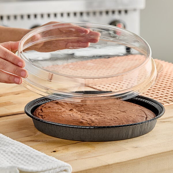 A person holding a clear lid over a brown cake in a pan.