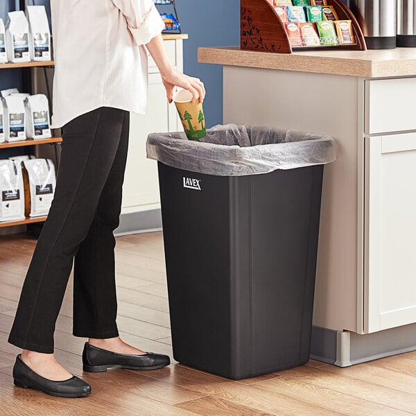 A woman in black pants putting a cup into a black Lavex square trash can.