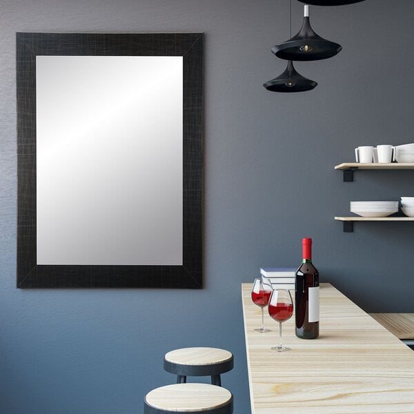 A BrandtWorks large black mirror mounted on a wall above a table with a bottle of wine and two glasses.
