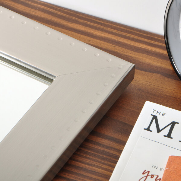 A BrandtWorks silver embossed mirror hanging above a table with a clock and picture frame.