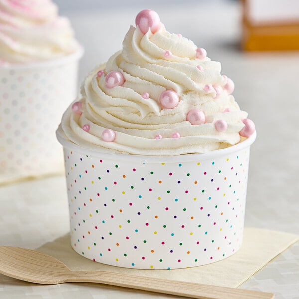 A white polka dot paper food cup filled with ice cream with pink and white frosting and pink beads on a table.