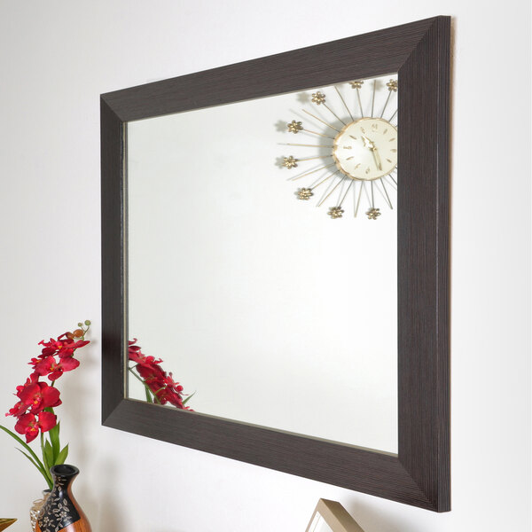 A BrandtWorks modern espresso finish mirror on a wall above a vase with red flowers with a clock with gold hands.