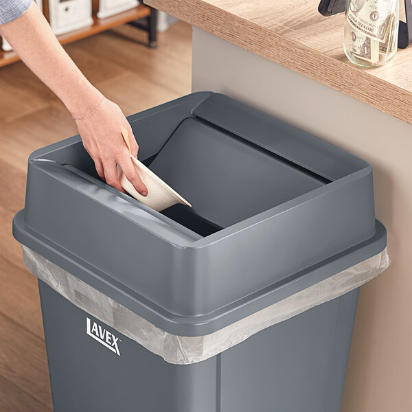 A person putting a paper bowl in a Lavex gray square trash can with a swing lid.