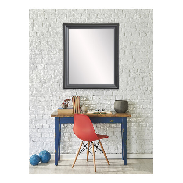 A BrandtWorks matte black mirror hanging on a white wall above a red chair with wooden legs.