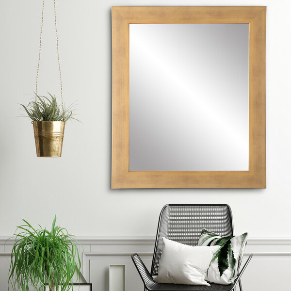 A BrandtWorks brushed gold mirror on a white wall above a chair with pillows and a plant.