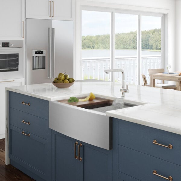 A Ruvati stainless steel farmhouse sink in a kitchen with blue cabinets and a white counter.