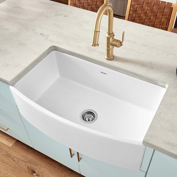 A white Ruvati Fiamma farmhouse sink with a gold faucet.