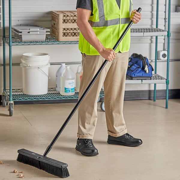 A man in a safety vest using a Lavex polypropylene push broom with flagged bristles and a metal handle to sweep a grocery store aisle.