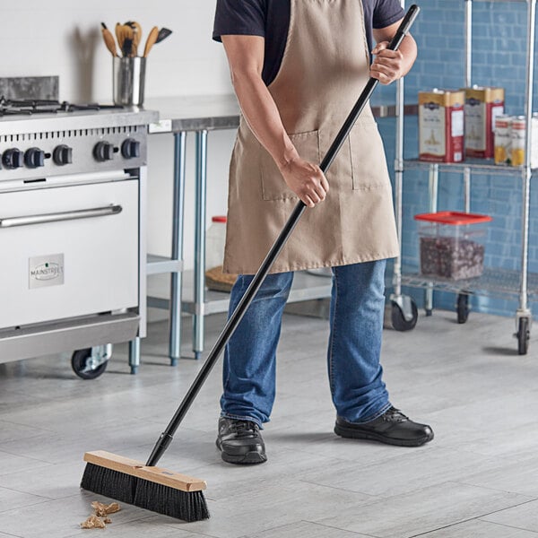 A man in an apron using a Lavex wood push broom to sweep the floor in a professional kitchen.