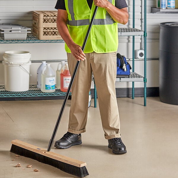 A man using a Lavex wood push broom with flagged bristles and a metal handle to sweep a floor in a grocery store aisle.