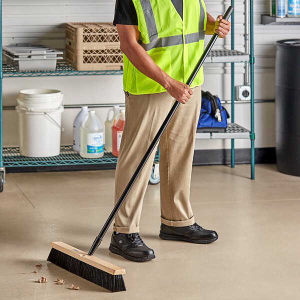 A man in a safety vest using a Lavex wood push broom with unflagged bristles to sweep a grocery store aisle.