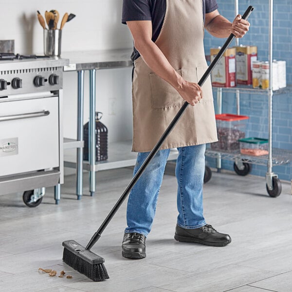 A man in an apron sweeping the floor with a Lavex Polypropylene Push Broom.
