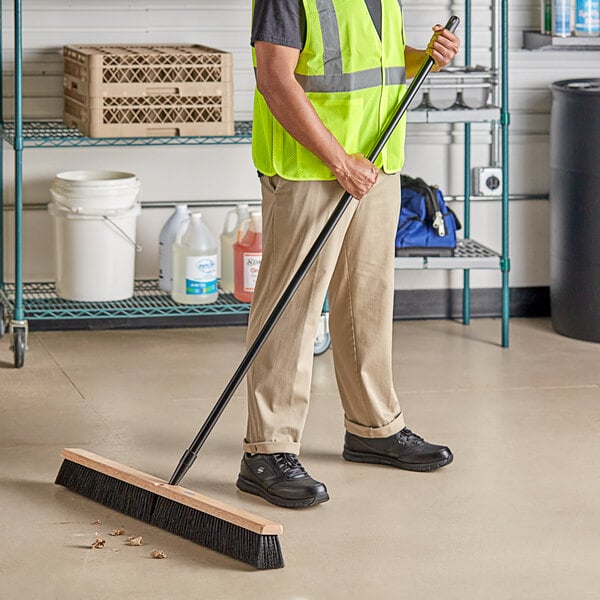 A man in a safety vest sweeping the floor with a Lavex wood push broom.