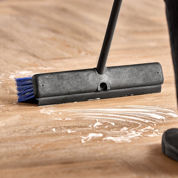 A push broom with unflagged bristles, a squeegee, and a long metal handle being used to clean a floor.