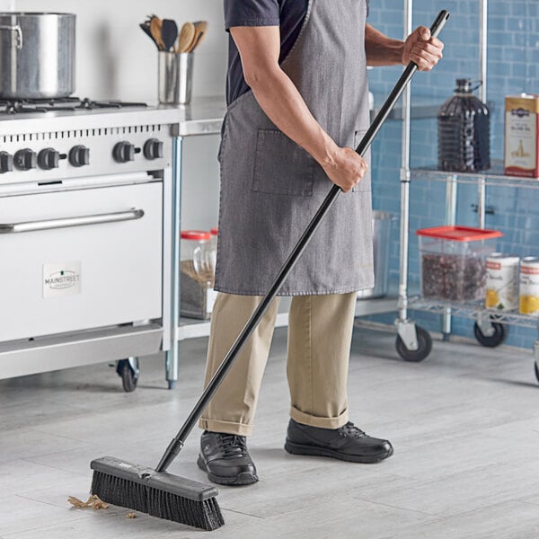 A man in an apron sweeping the floor with a Lavex 18" Polypropylene Push Broom.