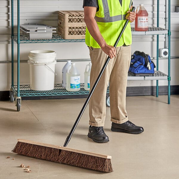 A man in a safety vest sweeping the floor with a Lavex wood push broom.