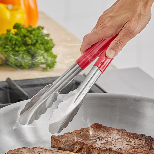 A hand holding Fourté red coated handle stainless steel scalloped tongs over a pan of steak.