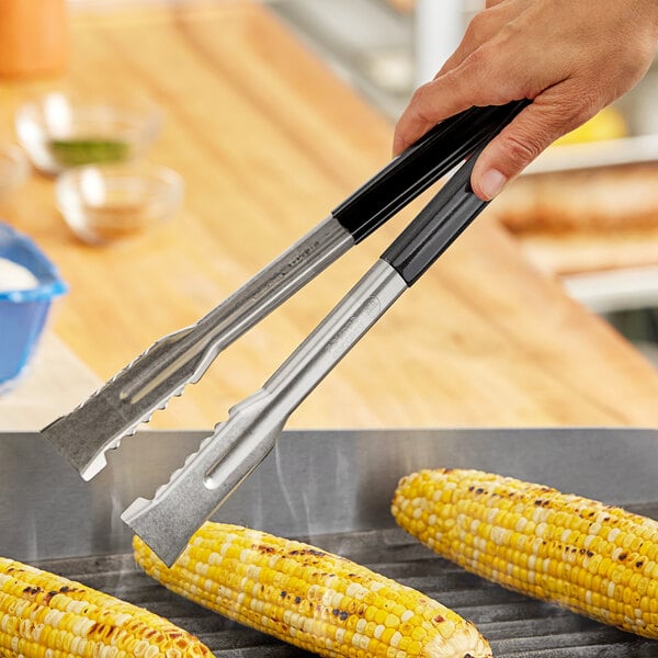 A person using Fourté black stainless steel grill tongs to grill corn on the cob.