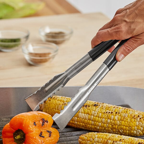 A person using Fourté stainless steel tongs to hold a corn on the cob over a grill.