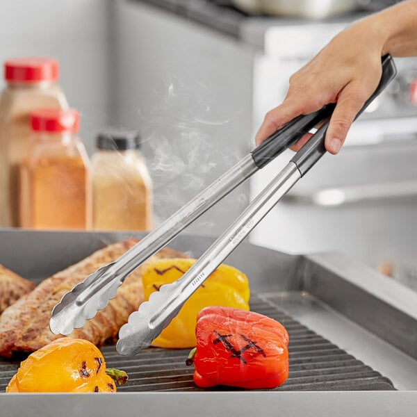 A person using Fourté black stainless steel scalloped tongs to cook vegetables on a grill.