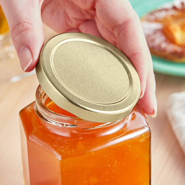 A person holding a 63 Lug gold metal lid on a jar of orange liquid.