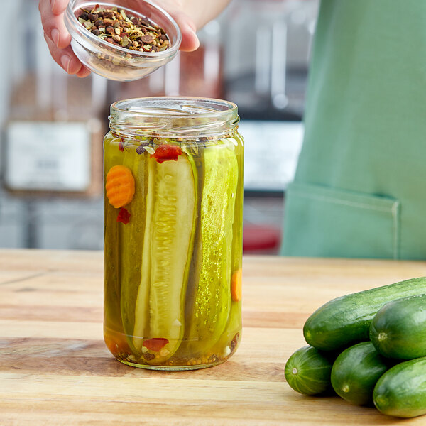 A person pouring spices into a jar of pickles on a counter.