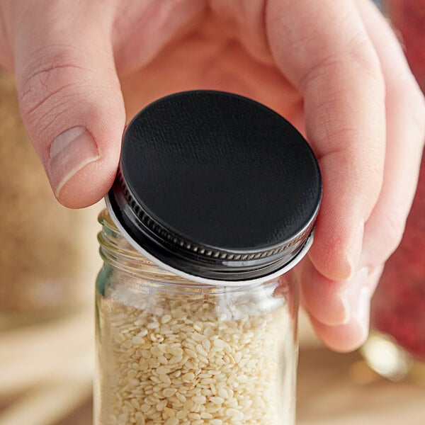 A hand holding a jar of sesame seeds with a black metal lid.