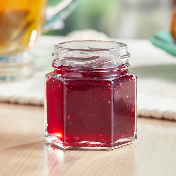 A 1.5 oz. hex glass jar of red liquid sitting on a table next to a glass of juice.