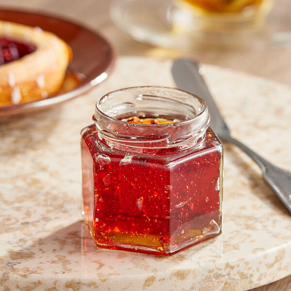 A 4 oz. hex glass jar of jam on a table next to a knife.