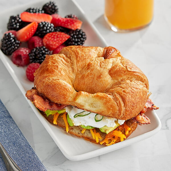 A Bridor fully baked sliced butter croissant sandwich with cheese and bacon on a white plate with berries and orange juice.